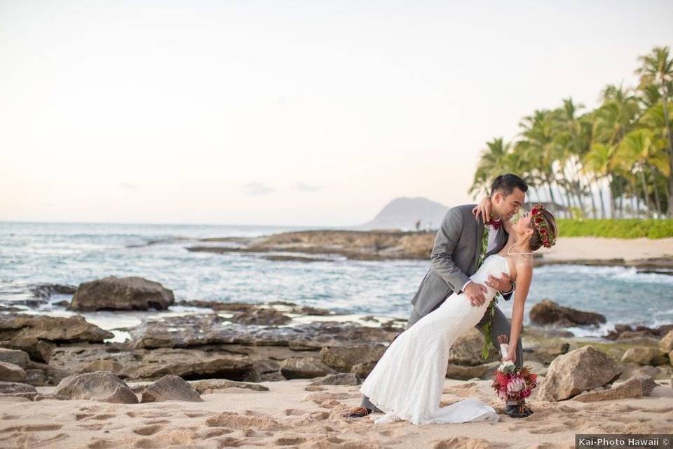 Hawaiian bride and groom standing on rocks along the ocean at sunset
