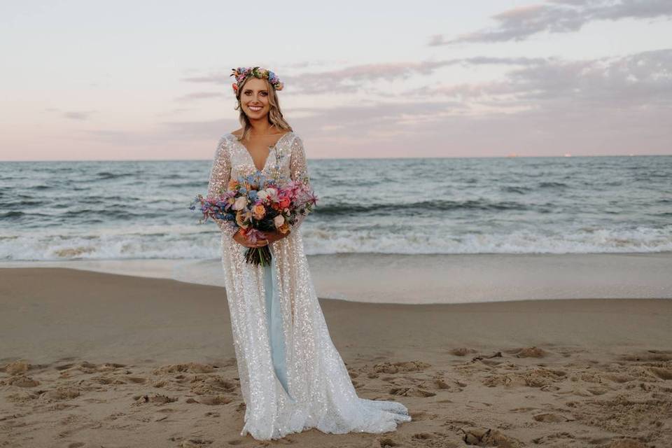 bride stands on the beach with the ocean in the background while holding a colorful bouquet and wearing matching flower crown