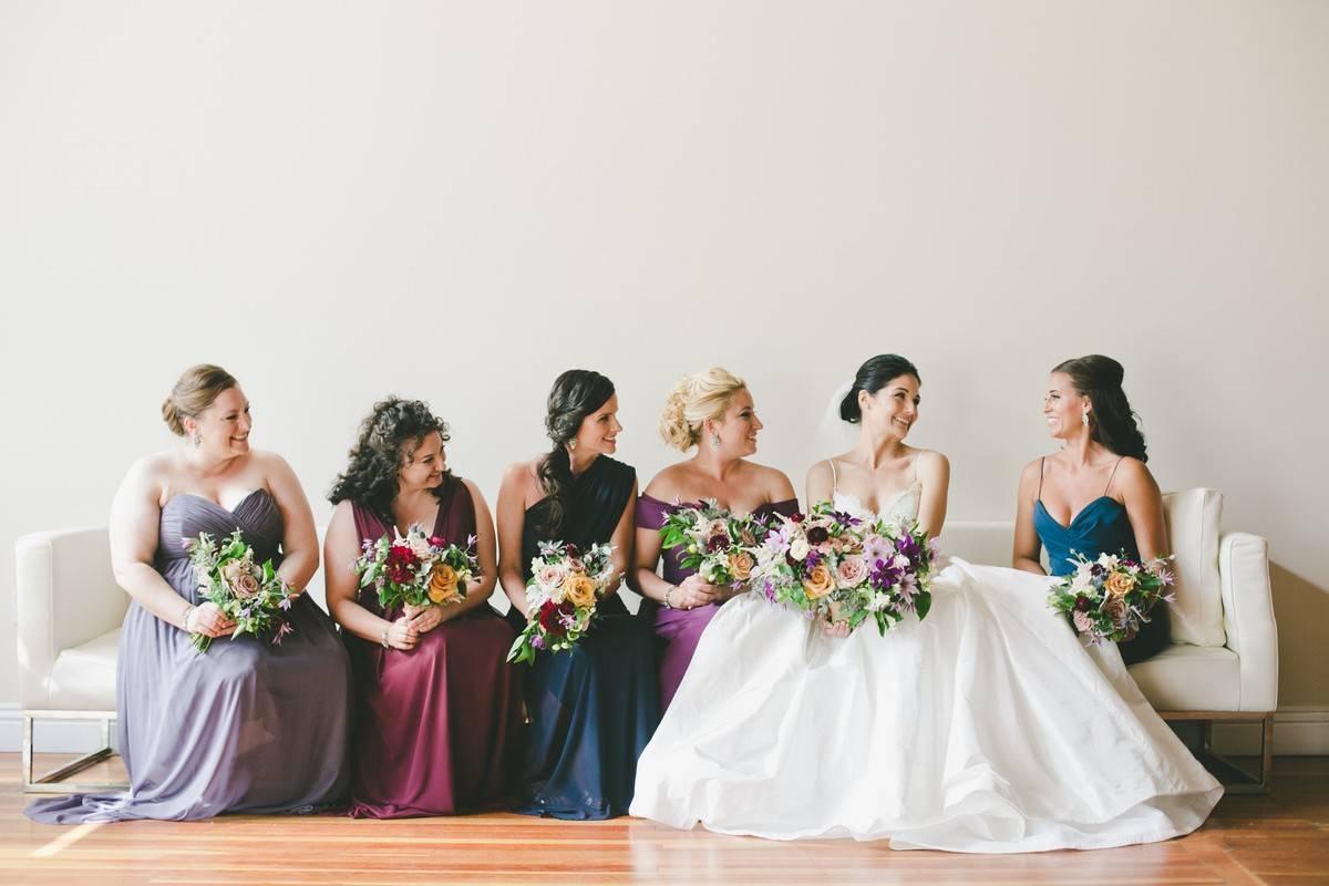 Bride sitting down on long sofa with bridesmaids wearing varying shades of jewel tones