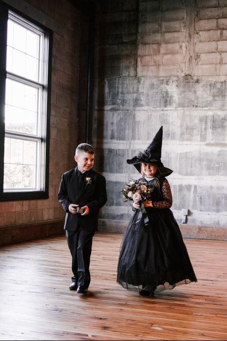 flower girl wearing black tulle dress and witch hat walks down the aisle with ring bearer