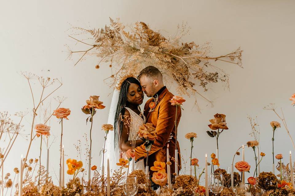 bride and groom stand behind table of modern flower arrangement with long-stem orange roses and taper candles