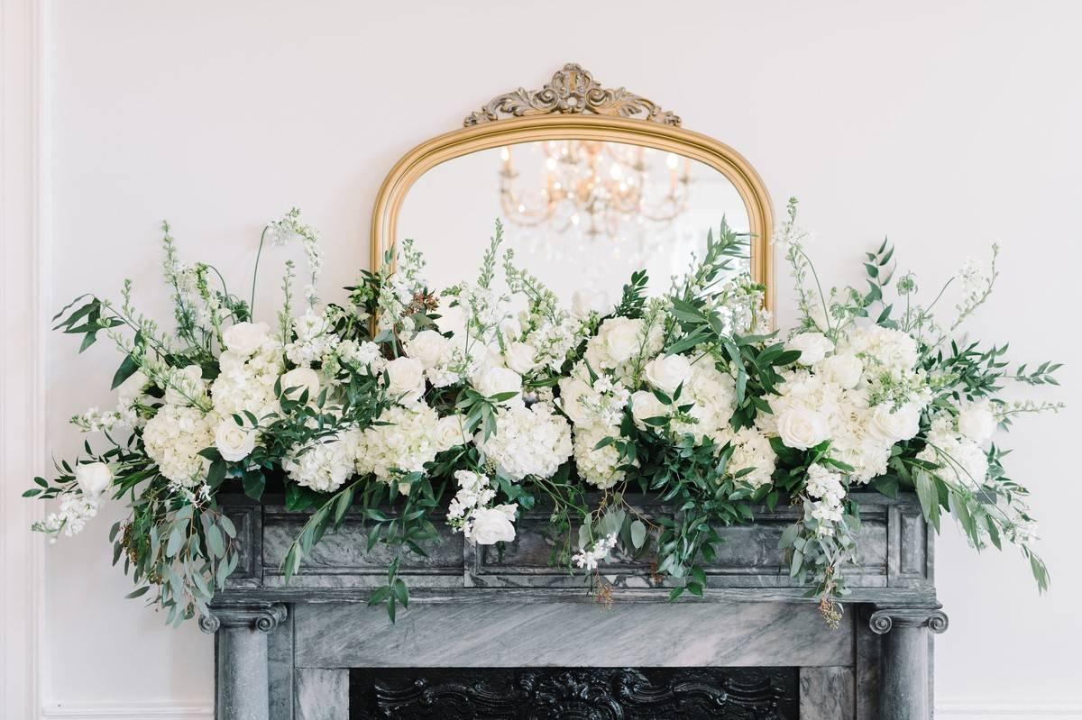 gray marble fireplace mantel is decorated with white flowers and greenery in front of antique gold frame mirror