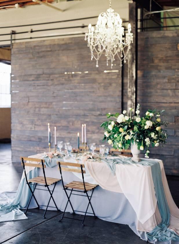 romantic wedding table setup with white tablecloth, light blue table runner, blue taper candles in gold candle sticks, and oversized greenery centerpiece