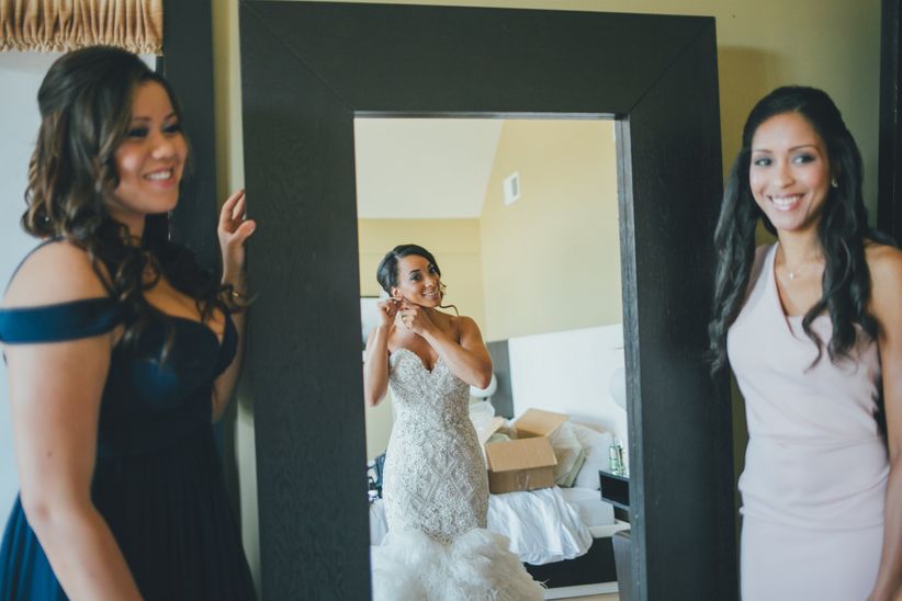 Cómo lucir bien en fotos de boda Novia mirando en el espejo mientras se prepara para su boda con dos damas de honor observando
