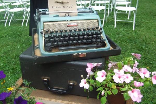 A Vintage Typewriter holds the Typed Program for the Ceremony, and is sitting atop a stack of old Bee-keeping boxes, surrounded by wildflowers. Beautiful! all items for rent.