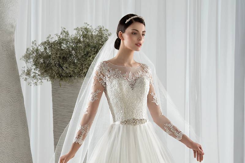 Style 582 <br> This classic tulle, Aline gown with a jeweled belt features delicate beaded lace appliques throughout the bodice, sheer neckline and long sheer sleeves.   The embellished sheer back features button closures and is finished with a Chapel length train.