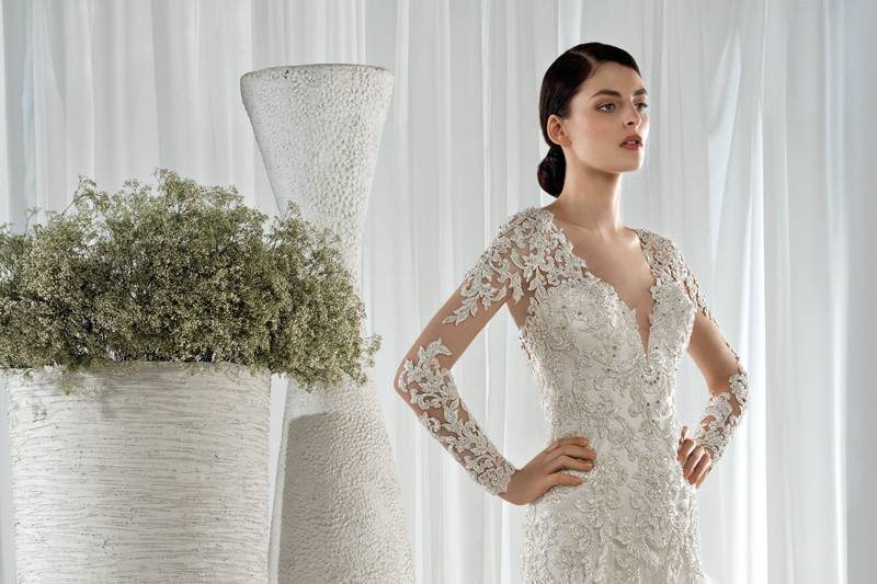 Style 616 <br> This striking Fit-N-Flare gown with plunging V-neckline is embellished with beaded lace on the bodice, long sheer sleeves and dramatic natural sheer back with button closure.  The multi-tiered Tulle skirt flows into a Chapel length train.