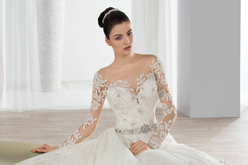 Style 648 <br> This romantic beaded lace Ball Gown features an illusion bateau neckline with sheer embellished long sleeves, and beaded waist- line. The back features an open sheer back with beaded lace applique embellished with button closures and a Chapel train.