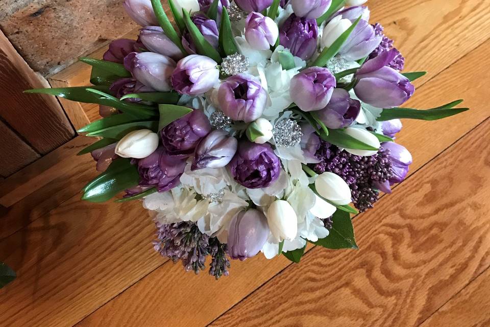 Stunning spring clutch bouquet of purple, lavender, and white tulips, with fragrant purple lilacs, and white hydrangea.