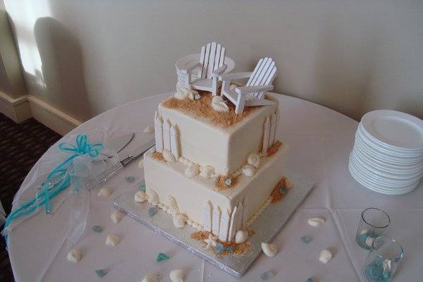 White square buttercream beach cake with gumpaste fencing, white chocolate shells and keepsake beach chairs.  Accented with teal candies and sea glass.