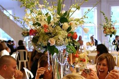 This elaborate display includes our tall glass pilsner with submerged tulip with abundant arrangement of green hydrangea, coral and ivory roses, white orchids, curly willow, bells of ireland and camelia foliage.  It is surrounded by hand-tied bouquets in footed vases with our large cylinder votives of water and lily grass sparkling by candlelight with our up lights for additional ambiance.  And we topped off the entire arrangement with crystal chandelier teardrops to give it that over the top elegance!