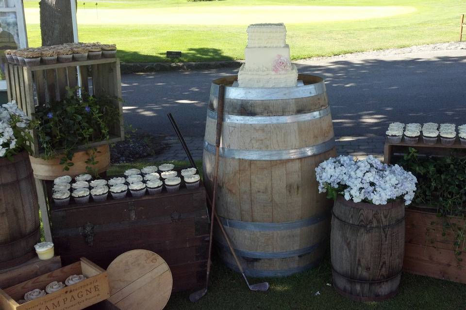 3 tier buttercream white wedding cake adorned with a single sugar rose in pale pink sitting atop a wine barrel.  Side crates hold cute rustic wedding cupcakes in several flavors.