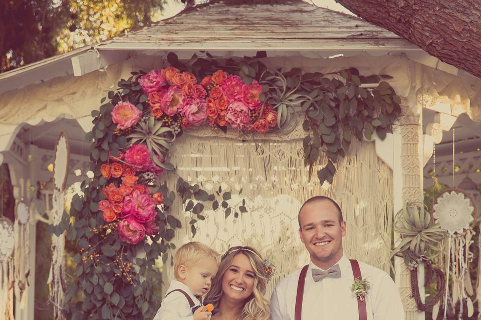 Bride and groom with their nephews. They're standing in front of the gazebo affixed with a macrame backdrop, the bride's dream catchers, and an arch piece we designed and assembled day-of.