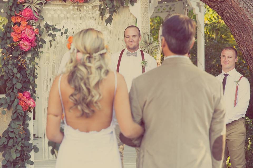 Father of the bride walking her down the aisle. She wears a small floral headdress while the groom and best man wear small boutonnieres of succulents and greenery. They are all in front of the gazebo at Green Gables Estate in San Marcos, CA, which we decorated with the bride's macrame backdrop, her dream catchers, and an arch piece with a lot of greenery.