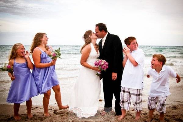 bride and groom kissing while children make silly faces at them, on the beach