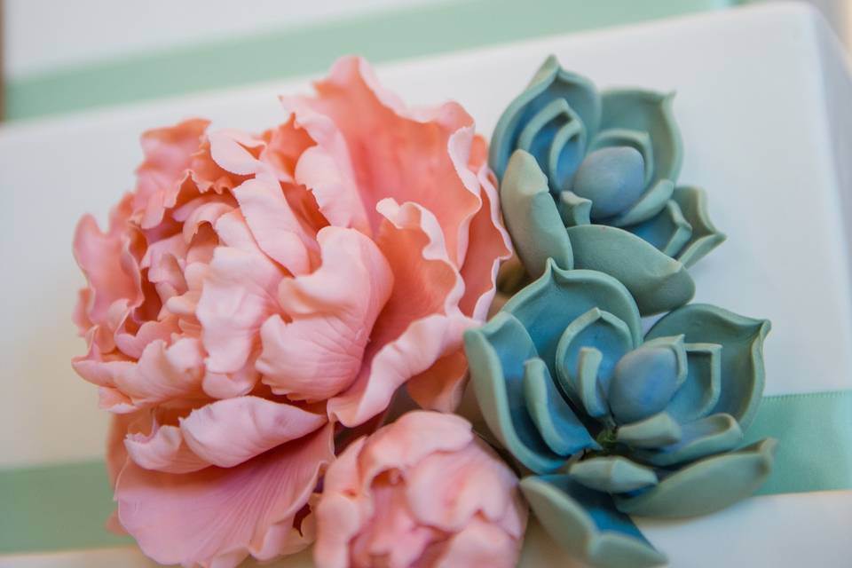 Closed peony, peony bud, and succulents handcrafted from sugar, to adorn a four-tiered square cake with mint green ribbon trim.