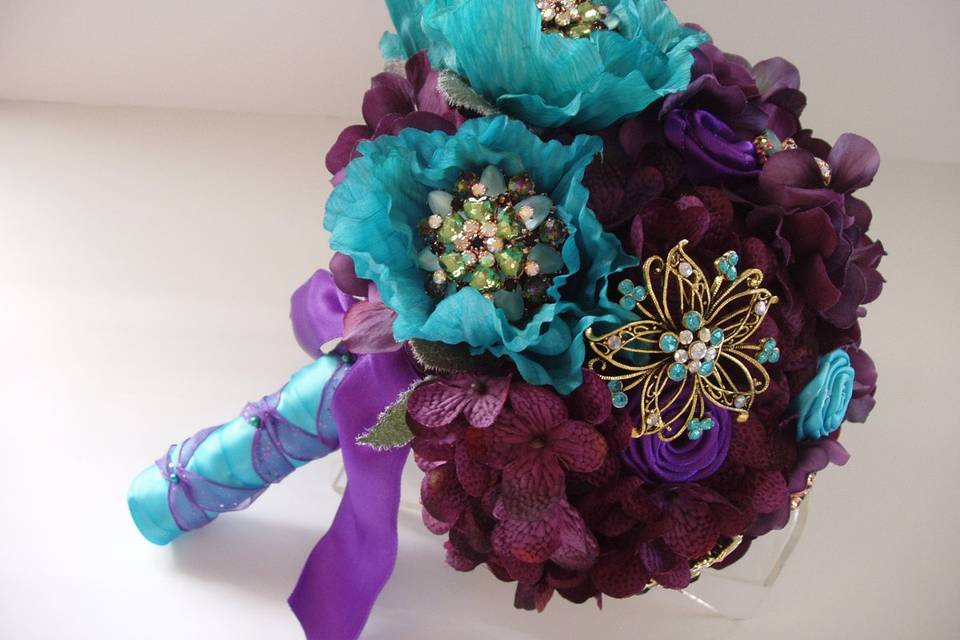Lovely and romantic quality silk bouquet created with eggplant hydrangea, turquoise poppy flowers, turquoise and purple satin ribbon roses. Turquoise rhinestone and crystal brooches are interspersed throuout the bouquet. The handle is wrapped in an turquoise satin ribbon with a purple ribbon criss cross. This bouquet is available in other colors.