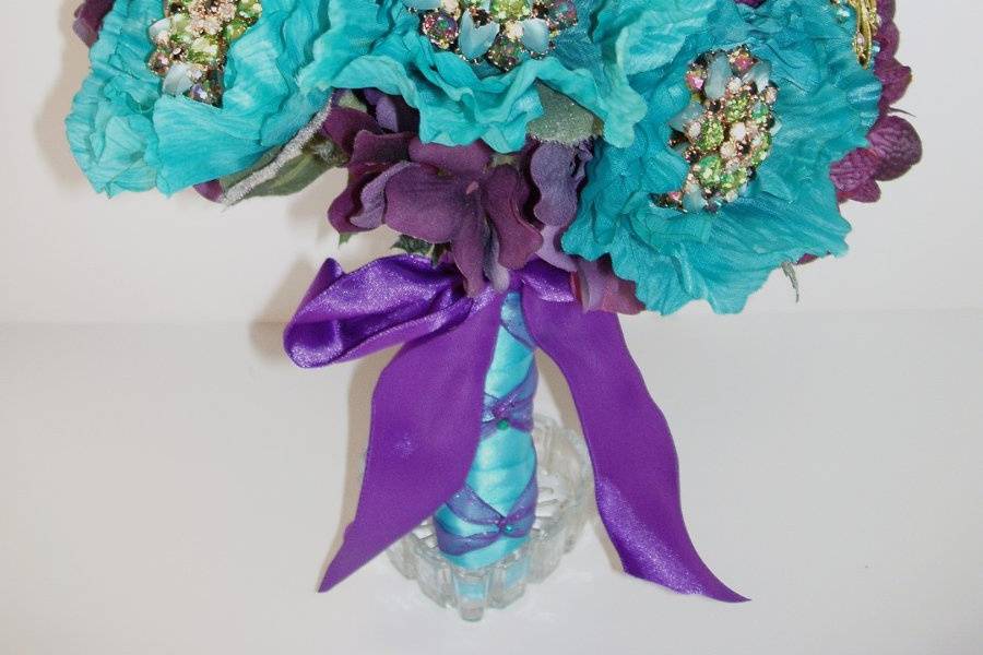 Lovely and romantic quality silk bouquet created with eggplant hydrangea, turquoise poppy flowers, turquoise and purple satin ribbon roses. Turquoise rhinestone and crystal brooches are interspersed throuout the bouquet. The handle is wrapped in an turquoise satin ribbon with a purple ribbon criss cross. This bouquet is available in other colors.