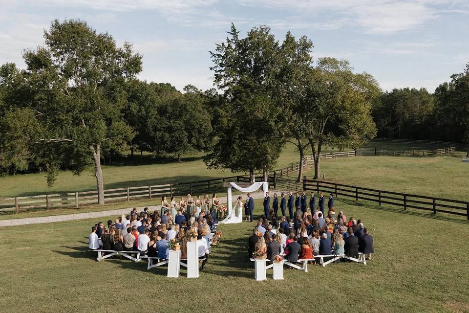 An outdoor ceremony on the fenced lawn, guests seated on white benches beneath the oaks