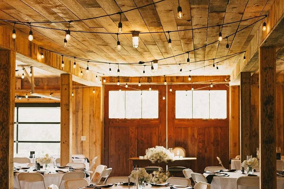 Round tables set for a reception inside the barn under warm string lights
