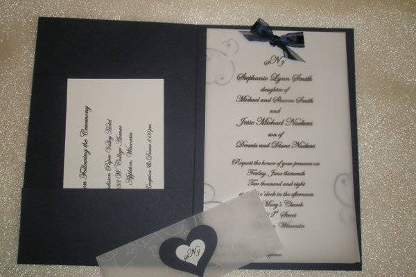 A navy pocket invitation featuring a bright white cardstock covered with a shimery vellum sheet.  Invitation is wrapped with a printed vellum band with heart-shaped monogram seal.