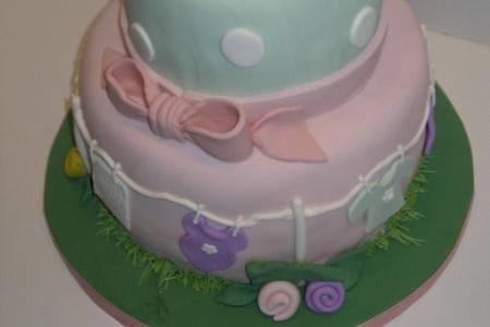 A baby shower cake covered in fondant with a clothesline of baby things blowing in the breeze. A baby on a rocking horse topper and butter cream grass circles the base tier.