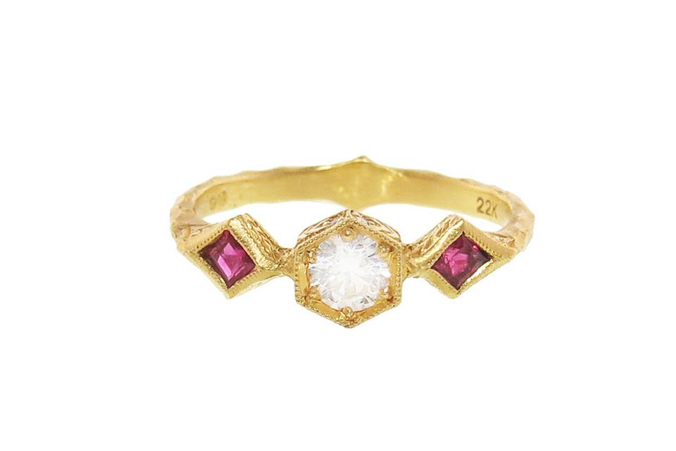 Indulge in a dazzling dose of resplendent color. Set in 22 karat gold, this ring features impressive .25 carat Burmese rubies that rest on either side of a .265 carat white diamond commanding attention as the focal point. The white diamond rests rests in a hand etched hexagon setting. Finished on a thin hammered band.
