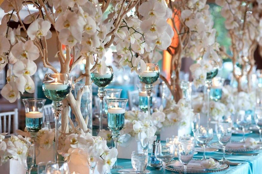 Beautiful blossoms in Winter White with exposed branches elevate this table setting for the Wells-Barrington wedding. With mineral water dyed in blue to add color towards the flowers, pick up the detail and design on the matching tablecloths.