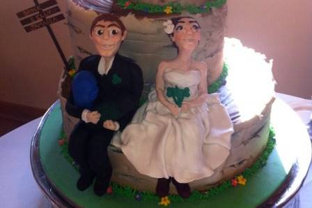 Nature-themed wedding cake with birch bark fondant, royal icing flowers, and hand-made clay sculptures of the couple. On the top is a tent and a small campfire, and on the side is a campground sign announcing where they're going on their honeymoon. The figures both have their hiking boots on, and he is holding their backpacks.