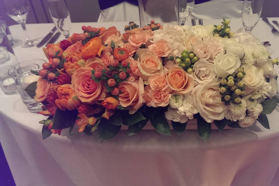 Low centerpiece for sweetheart table in pale shades of orange and white that came together to make an ombre arrangement.  It included roses, tulips, hypericum berries, ranunculus and greens.