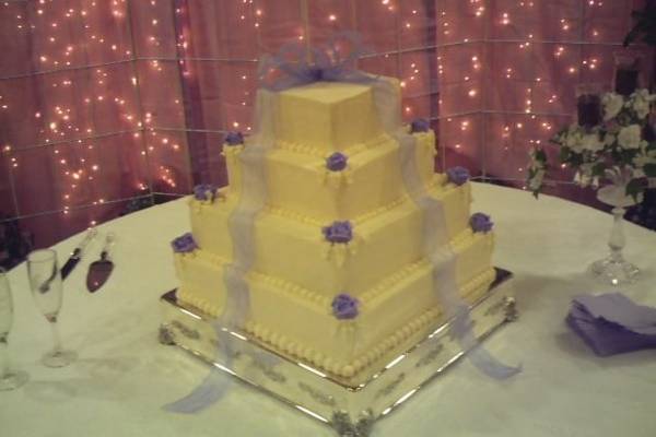 Four tiered pyramid cake with edible roses placed on each corner and a large bow placed as cake topper with the bow ties cascading down on each side. Wedding cake placed on sterling silver plateau.