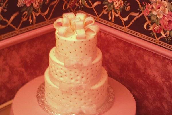 Three tiered traditional wedding cake covered with fondant with silver ribon around the bottom of each tier and edible silver balls randomly placed on each tier. Cake topper is a large bow made with fondant.