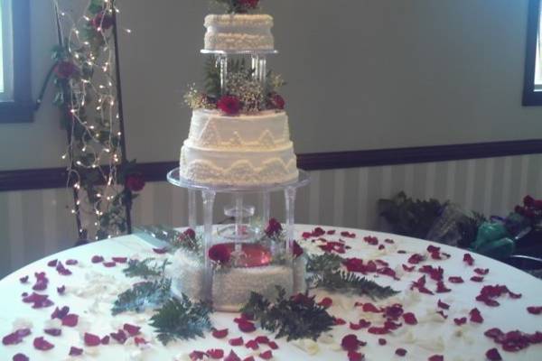 Four tier cake with two sets of columns and water fountain on top of bottom tier. Red rose bouquet as cake topper between columns and rose petals sprinkle on cake table.