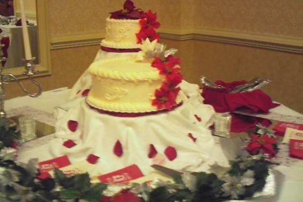 Two seperate cakes, with flowers cascading down on one side, placed atop multi-level stands. The stands are draped with a white tablecloth and flower petals sprinkle around the cake.