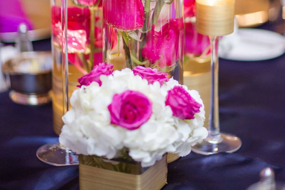Low arrangement with submerged magenta roses in a set of 3 cylinder vases. Next to 2 small cubes with white hydrangeas and set of 3 Halo stem candle holders with floating candles.