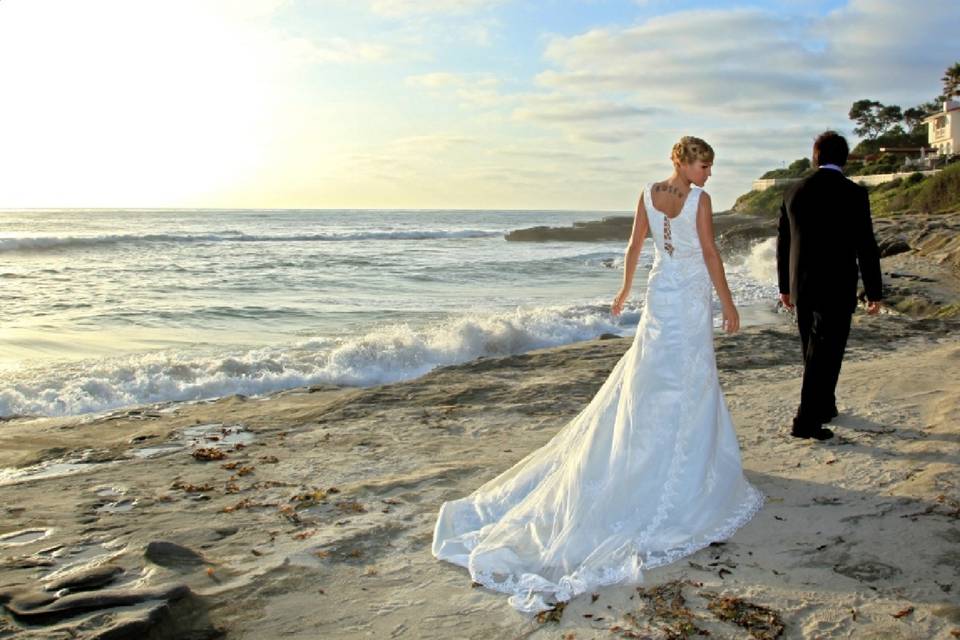 Couple walking along beach with blue and yellow sky. Bride's dress is lovely and long, sweeping across sand