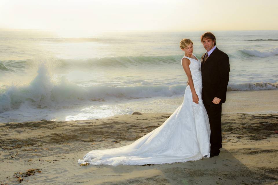 Bride with her groom standing on beach with wave splashing in background. Overall photos has a painting effect. La Jolla, CA.