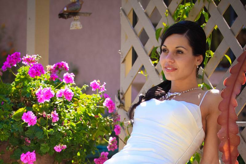 Bride in white dress sitting on garden bench with flowers and lattice around her.