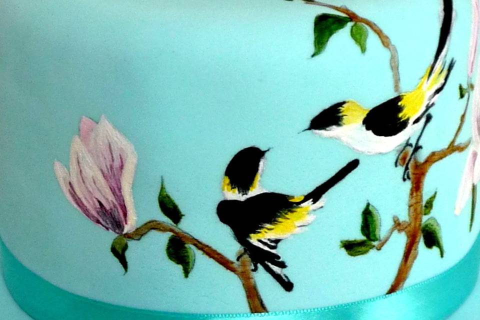 A pair of handpainted cocobutter birds perch on Magnolia blossoms.