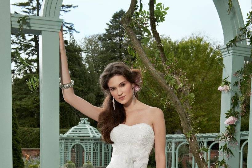 Style No. 42426_1387 <br>Features include a square neckline with decadent lace bodice and beautifully textured organza trumpet skirt with belted side flower detail that is bejeweled with a pearl and rhinestone center, and ending with a chapel train. Words alone cannot do this wedding gown the justice it truly deserves.