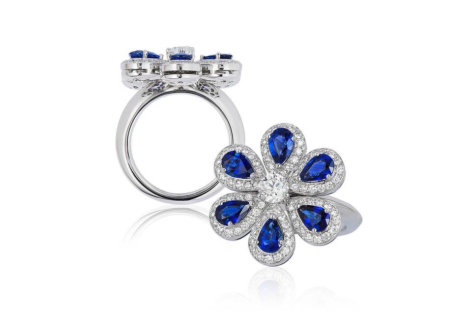 Blue Sapphire and Diamond RingThis 18k white gold ring features a .45 carat center diamond surrounded by six .50 carat blue sapphires in a beautiful blossoming flower pattern. Finished with beautiful micro pave around each blue sapphire, this statement ring will be the center of any occasion you wear it to.