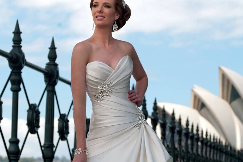 Y21263 <br>
A majestic ball gown in Duchess satin showcases the unparalleled craftsmanship we have all come to admire in the Sophia Tolli label. The exceptional fit of the bodice is enhanced with classic draping and crystal brooches, while folds of luxuriant satin create a sculptural effect in the skirt and chapel length train. Susan offers a functional corset back and removable straps, completing this remarkable gown.