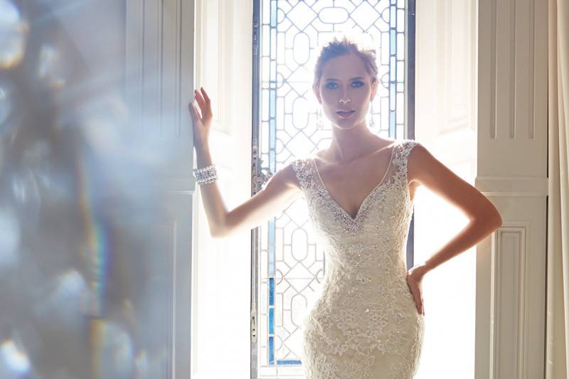Y21432 - Leigh<br>
2014 Collection – Enhance your beauty and embrace your inner goddess with Leigh, a slim sheath in point d’esprit and lace appliqués. Crystal hand-beading frames the plunging V-neckline and sheer cap sleeves while lace appliqués and scattered beading highlight the sheer back bodice complete with cascading crystal buttons that conceal a zipper closure. A scalloped chapel length train finishes off this stunning gown.