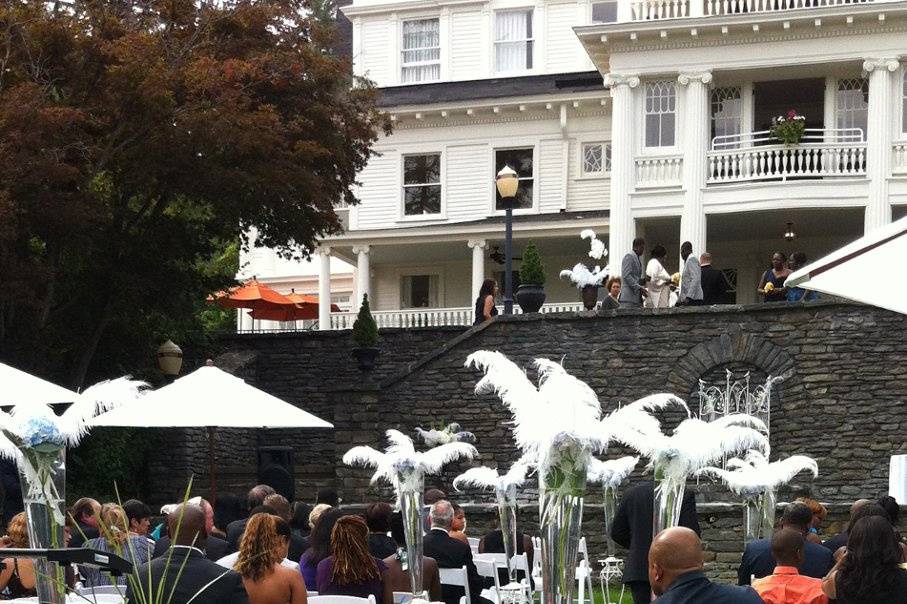 Tall glass vases holding Hydrangeas and plumed with ostrich feathers sit atop white wrought irons stands flanking the aisle at The Mansion at Noble Lane in Bethany, PA.