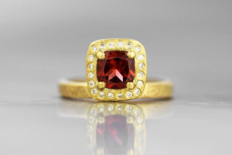 Custom created cushion cut garnet with white diamond halo, offset on a textured 18k yellow gold band.