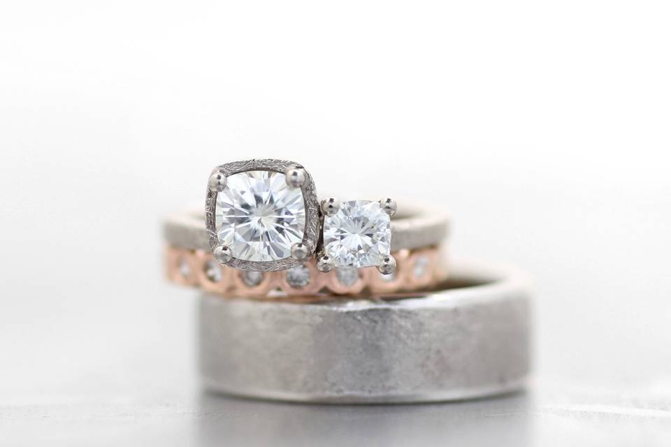 14k white gold engagement ring with cushion cut moissanite, rose gold wedding band with white diamonds, and white gold men's band.