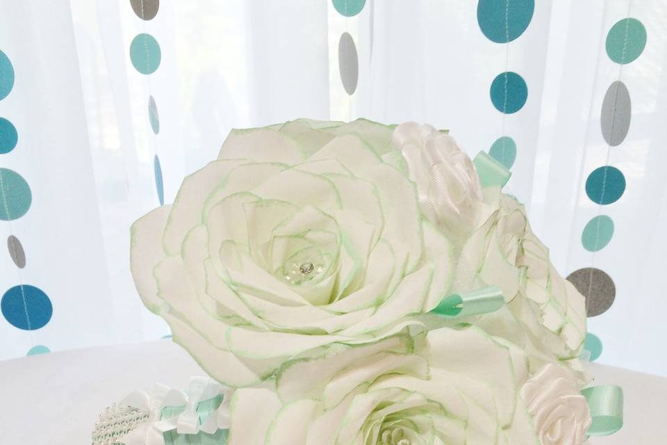 Handmade coffee filter Roses and Rose buds with the tips of the flowers in mint green and a small acrylic and rhinestone flower in the middle of each rose. There are white foam roses and white tulle mixed in with the flowers. Around the stems is mint green satin ribbon and white rhinestone ribbon.