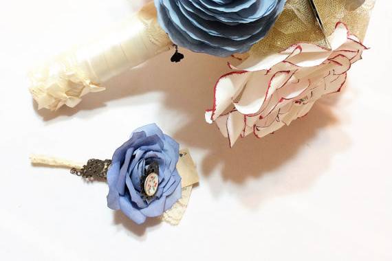 Alice in Wonderland themed wedding bouquet. There are smokey blue, gold and red tipped paper Roses and Peonies surrounded by gold glittered tulle. Small playing cards, tiny tea cups and saucers and charms accent the bouquet. The handle is wrapped in satin ribbon, lace and bows.