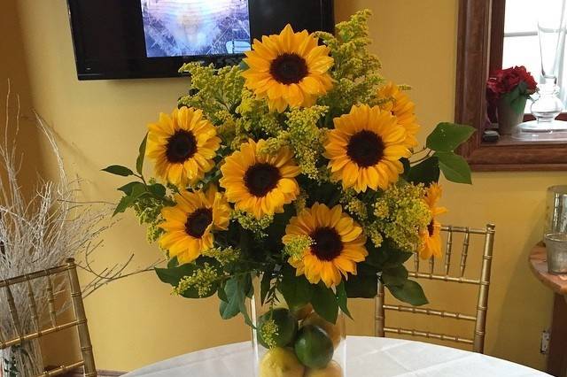 Fresh simple arrangement designed with sunflowers ,solidago and lemon leaf on top of cylinder vase filled with lemon and limes .