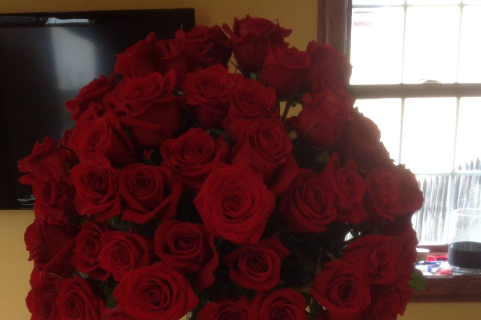 Lush ball of all red roses wedding arrangement on top of cylinder vase accented with a diamond band around center .